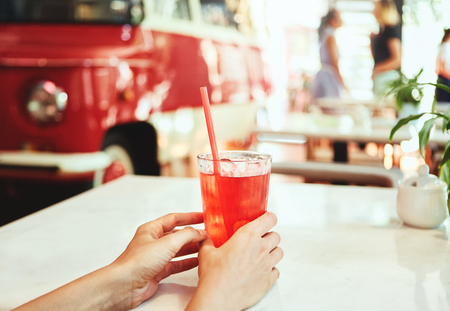 Woman drinking red lemonade or cocktail in cafe. Hands holding a cocktail on the foreground. Young female enjoying a glass of soft drink with a straw sitting at a restaurant table. Summertime concept. Side view.の写真素材