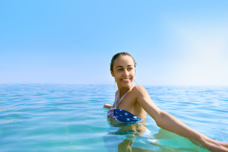 Portrait of young beautiful smiling woman with tanned body in the water on tropical sunny beach. Slim pretty model is swimming in the sea with blue sky background. Summer vacation and tanning concept.の写真素材