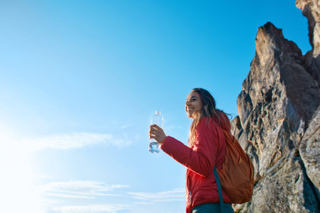 woman hiker with backpack, wearing in red jacket, standing on edge of cliff against a blue sky...