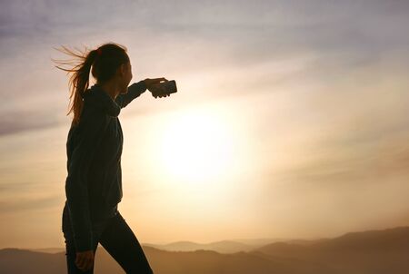 Silhouette of young woman standing on edge of mountain and and taking a photo on sunset sky and mountains background. Travel and active lifestyle concept.の写真素材