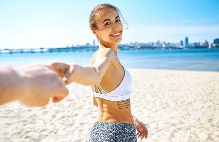 Portrait of beautiful sexy happy girl on the sand city beach with sea background. Young smiling woman having fun and and holding someone hand at the coast against blue sky and sea. Summertime and follow me concept.の写真素材