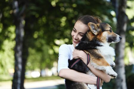 Welsh Corgi Pembroke dog and smiling happy woman together in a park outdoors. Young female in white shirt embracing, holding a her cute pet. Concept friendship with dog and human, cute moments, happines.の写真素材