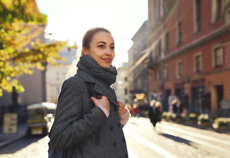 Young attractive woman in a warm scarf and gray coat walking city street. Stylish smiling girl posing at camera, enjoying a sunny autumn day.の写真素材