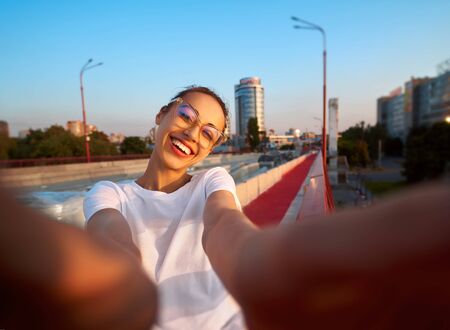 Bright summer lifestyle portrait of young pretty woman in eyewear, red skirt and white T-shirt, taking a selfie on camera, standing on the bright red bridge at evening with city background. Trendy hipster girl smiling, looking into camera, enjoying warm summer day on sunset. Bright color, summertime and lifestyle concept.の写真素材