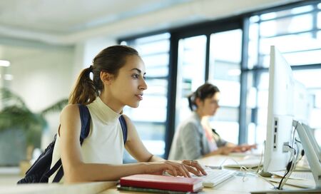 students in computer class, or library. Beautiful happy young woman with a backpack and notebooks inside a college campus using computer. Young female university student with books in campus. Education, knowledge concept, copy space.の写真素材