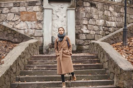 Autumn outdoor portrait of beautiful young woman walking in autumn park, goes down the stairs, wearing beige coat and warm knitted scarf. Colorful fallen leaves and trees on background. Season and people concept.の写真素材