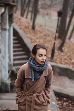 Autumn outdoor portrait of beautiful young woman walking in autumn park, wearing beige coat and warm knitted scarf. Colorful fallen leaves and trees on background. Season and people concept.の写真素材