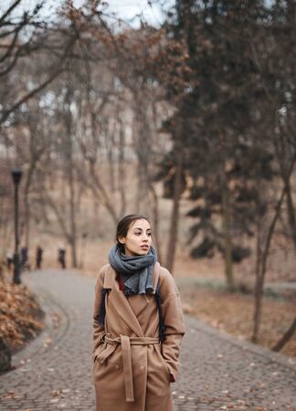 Autumn outdoor portrait of beautiful young woman walking in autumn park, wearing beige coat and warm knitted scarf. Colorful fallen leaves and trees on background. Season and people concept.の写真素材