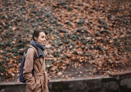 Autumn outdoor portrait of beautiful young woman walking in autumn park, wearing beige coat and warm knitted scarf. Colorful fallen leaves on background. Season and people concept.の写真素材