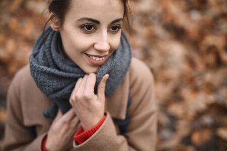 Close up autumn outdoor portrait of beautiful smiling woman in autumn park, wearing beige coat, wraps in warm knitted scarf. Season and people concept.の写真素材