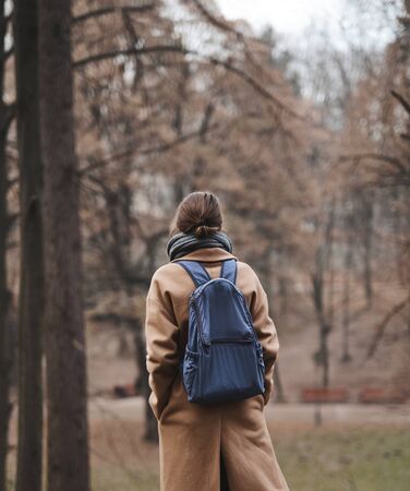 Autumn outdoor portrait of beautiful young woman walking in autumn park and enjoying the fall landscape, wearing beige coat and warm knitted scarf. Colorful fallen leaves and trees on background. Season and people concept. Back vievの写真素材