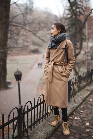 Autumn outdoor portrait of beautiful young woman walking in autumn park and enjoying the fall landscape, wearing beige coat and warm knitted scarf. Colorful fallen leaves and trees on background. Season and people concept.の写真素材