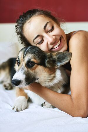 Portrait of happy smiling woman with her cute Welsh Corgi dog lying on the bed at morning, morning greeting. Beautiful brunette female hugging funny puppy, has good relationships with pet.の写真素材