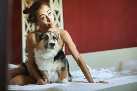Young woman with her cute Welsh Corgi dog on the bed in bedroom at morning, makes grimace, having fun.の写真素材