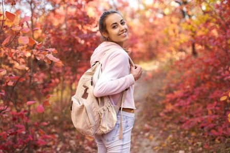 Autumn woman turns around at camera with charming smile. Happy smiling young woman in hoody and with backpack walking in autumn park with colorful leaves on fall nature background, in good mood. Outdoor atmospheric lifestyle portrait. Season and people concept.の写真素材