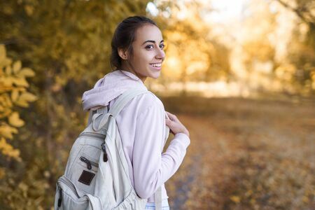 Autumn woman turns around at camera with charming smile. Happy smiling young woman in hoody and with backpack walking in autumn park with colorful leaves on fall nature background, in good mood. Outdoor atmospheric lifestyle portrait. Season and people concept.の写真素材