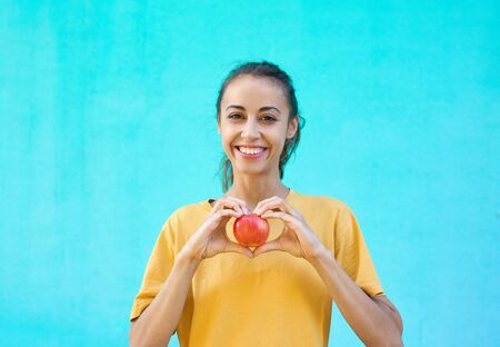 Pretty smiling girl holding apple and making a heart gesture with her fingers. Young brunette woman posing on a colorful bright cyan background.の写真素材