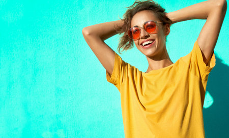 Fashionable cheerful young woman posing on blue background, wearing yellow t-short and trendy red eyeglasses. Happy girl has attractive look, posing to the camera and showing different emotions.の写真素材