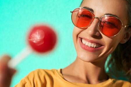 Close up portrait of a young stylish woman smiling at camera, showing big red lollipop against blue background. Cheerful girl wearing yellow t-short and trendy red eyeglasses.の写真素材