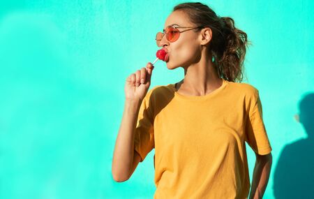 young stylish smiling woman standing against blue background and joyfully licking a big red lollipop. Cheerful girl wearing yellow t-short and trendy red eyeglasses.の写真素材