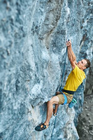 young strong man rock climber climbing on a high vertical limestone cliff, reaching holds, making hard wide move and gripping hold, attaching rope. Conquering, overcoming and active lifestyle concept.の写真素材