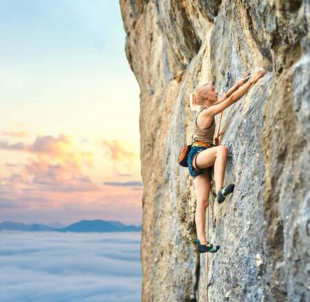 side view of young athletic woman rock climber climbing on the cliff. a woman climbs on a vertical rock wall with sunset sky on background. Conquering, overcoming and active lifestyle concept.の写真素材