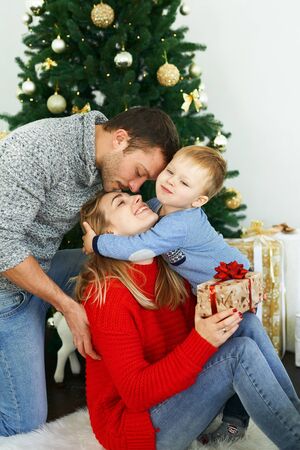 Young family celebrating Christmas at home, sitting near beautiful Christmas tree, laughing, embracing and giving presents. Young parents and their little son enjoying their holidays together. Merry Christmas and Happy New Year!の写真素材