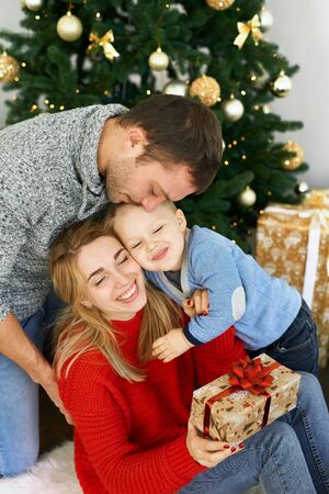 Young family celebrating Christmas at home, sitting near beautiful Christmas tree, laughing, embracing and giving presents. Young parents and their little son enjoying their holidays together. Merry Christmas and Happy New Year.の写真素材