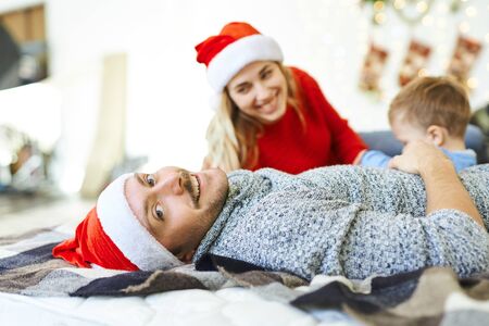 Portrait of handsome smiling man in red Santa cap, lying on the bed with little son and wife. Winter holiday, Christmas and New Year concept. happy time together.の写真素材