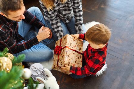 Young family celebrating Christmas at home, sitting near beautiful Christmas tree, laughing, embracing and giving presents. Young parents and their little son enjoying their holidays together. Merry Christmas and Happy New Year.の写真素材