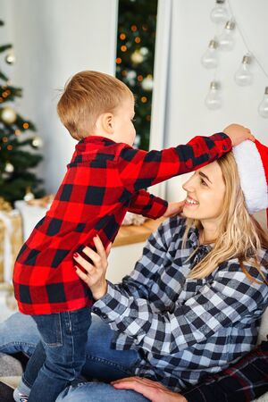 Little boy puts on a Christmas Santa hat to mom. Celebrating Christmas and New Year, having fun, happy time together.の写真素材