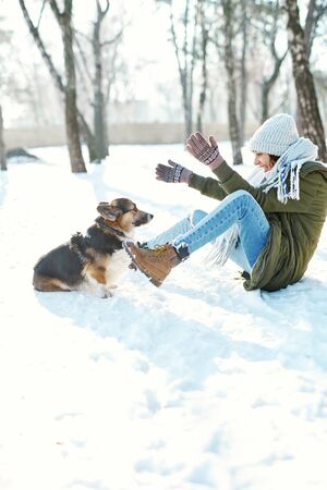 Young happy woman in woolen hat and long warm scarf playing with her pet in snowly winter park at frozzy sunny day. Happy time together, cute dog Welsh Corgi Pembroke, winter holodays.の写真素材