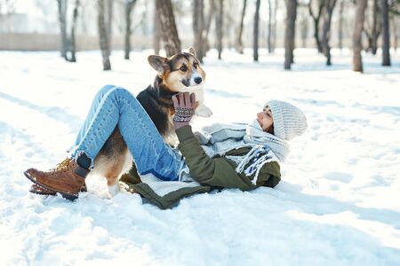 Young happy woman in woolen hat and long warm scarf playing with her pet in snowly winter park at frozzy sunny day. Happy time together, cute dog Welsh Corgi Pembroke, winter holodays.の写真素材