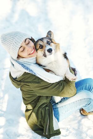Winter portrait of happy woman in woolen hat and long warm scarf holding, emrasing her pet in snowy winter park at frosty sunny day. Happy time together, cute dog Welsh Corgi Pembroke, winter holodays.の写真素材