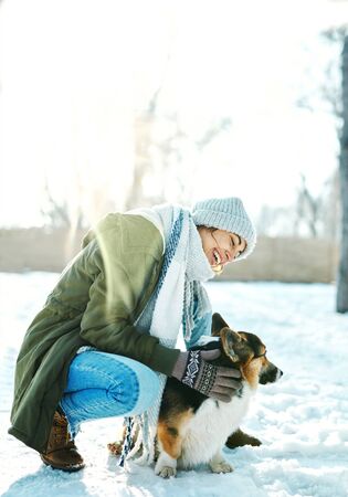 Young happy woman in woolen hat and long warm scarf stroking her pet and having fun in snowy winter park at frosty sunny day. Happy time together, cute dog Welsh Corgi Pembroke, winter holodays.の写真素材