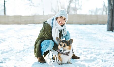 Young happy woman in woolen hat and long warm scarf smiling to camera, stroking her pet and having fun in snowy winter park at frosty sunny day. Happy time together, cute dog Welsh Corgi Pembroke, winter holodays.の写真素材