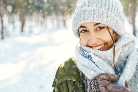 Closeup portrait of funny smiling woman in woolen hat and long warm scarf at snowy winter park in frozzy sunny day. Flying snowflakes. Happy winter time, having funの写真素材