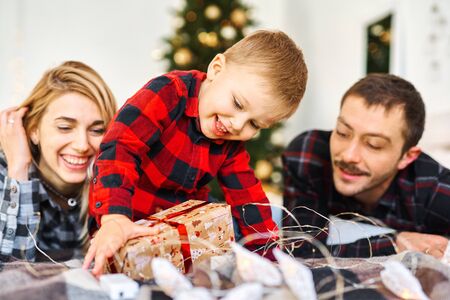 Young parents giving a Christmas present to little son at Chrismas morning in bed. Christmas family spending time together at home, Xmas tree on background. Merry Christmas and Happy New Year.の写真素材