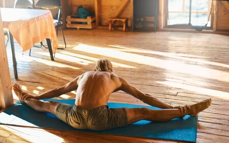 strong athletic handsome tanned man with naked torso doing stretching at home in wooden room at sunny morning. fitness, yoga, morning exercising, healthy lifestyle.の写真素材