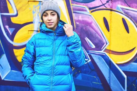 Portrait of young attractive woman wearing in blue down jacket and knitted hat posing against wall painted with graffiti. Urban clothing style, urban background.の写真素材