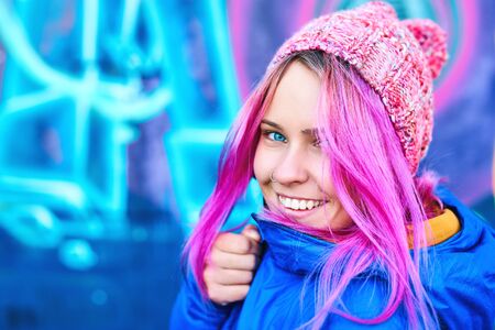 Closeup outdoor portrait of smiling funny woman in blue down jacket and pink hat posing against urban background. Urban clothing style, street style, lifestyle portrait.の写真素材