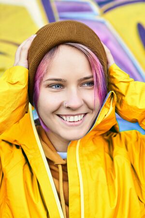 Urban bright portrait cool young woman with cheerful smile in yellow jacket and knitted hat posing against wall painted with graffiti. Urban clothing style, street style, lifestyle portrait.の写真素材
