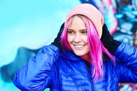 outdoor portrait of smiling funny woman in blue down jacket and pink hat posing against urban background. Urban clothing style, street style, lifestyle portrait.の写真素材