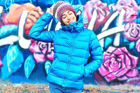 Portrait of young smiling woman wearing in blue down jacket and knitted warm hat posing against wall painted with graffiti. Urban clothing style, urban background.の写真素材