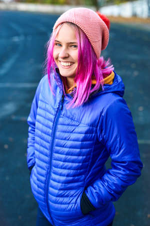 outdoor portrait of smiling funny woman in blue down jacket and pink hat posing against urban background. Urban clothing style, street style, lifestyle portrait.の写真素材