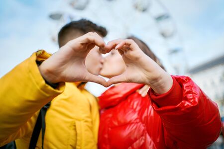 young romantic couple on date, embracing and kissing through a heart gesture made with their fingers, spending time together in the city. Lovers wearning in bright yellow and red down jackets. ferris wheel on background. Romantic, dating and Valentines Day concept. Focus on the fingersの写真素材
