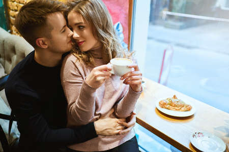 young Happy romantic couple sitting over the window in cafe, embracing, kissing and drinking coffee. boyfriend tenderly hugging girlfriend in cozy cafe. Romantic, love and Valentine's Day concept.の写真素材