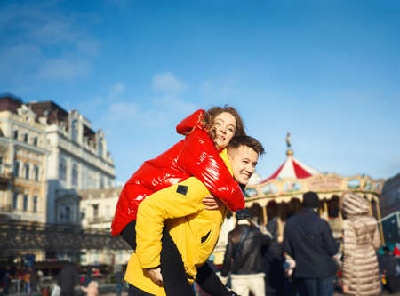 Happy young couple walking city street at bright sunny day, having fun together and looking happy. wearning in bright yellow and red down jackets. handsome young man carrying his girlfriend on shoulders. Romantic, love and Valentine's Day concept.の写真素材