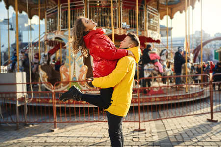 young romantic couple in love wearing in bright yellow and red down jackets walking city, hugging on french carousel background. Handsome man carrying young attractive woman on the hands.の写真素材