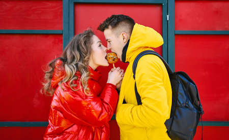Happy young winter couple in bright colorful warm clothes eating sweets outdoors on red background. beautiful couple in love walking city sunny day and enjoying together Valentines day holidayの写真素材